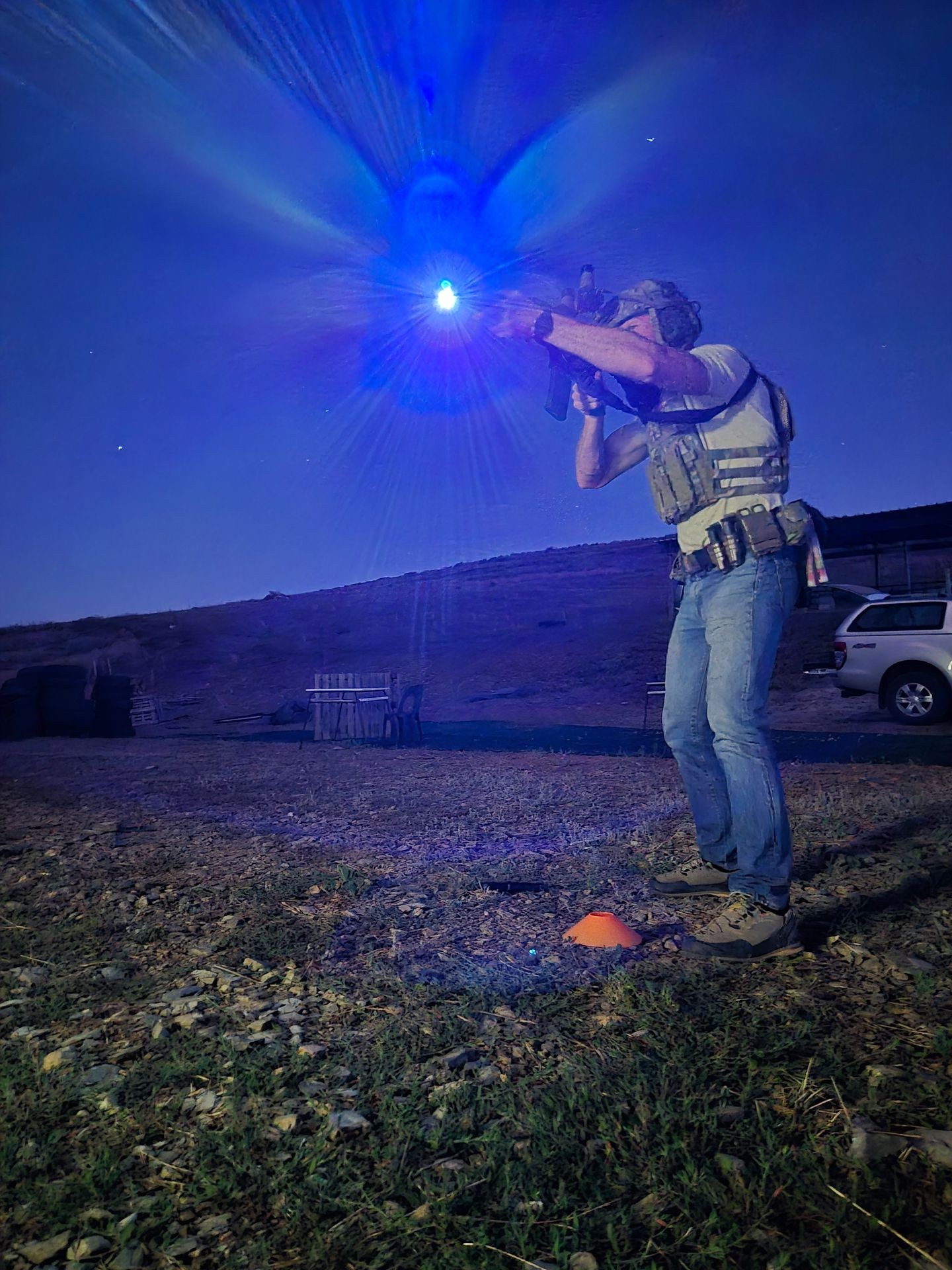Person in tactical gear aiming a rifle with a bright blue light at night on rocky terrain.