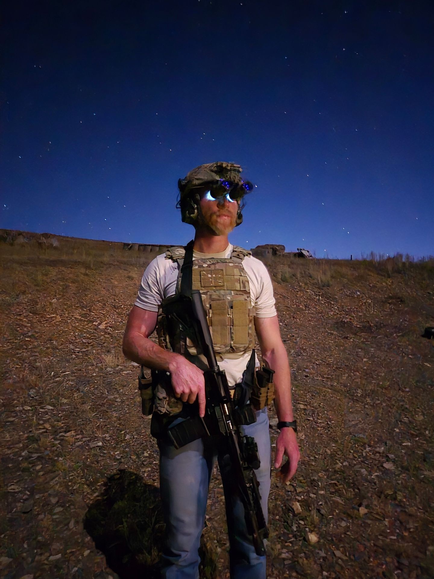 Person wearing tactical gear and night vision goggles, standing outdoors at night under a starry sky.