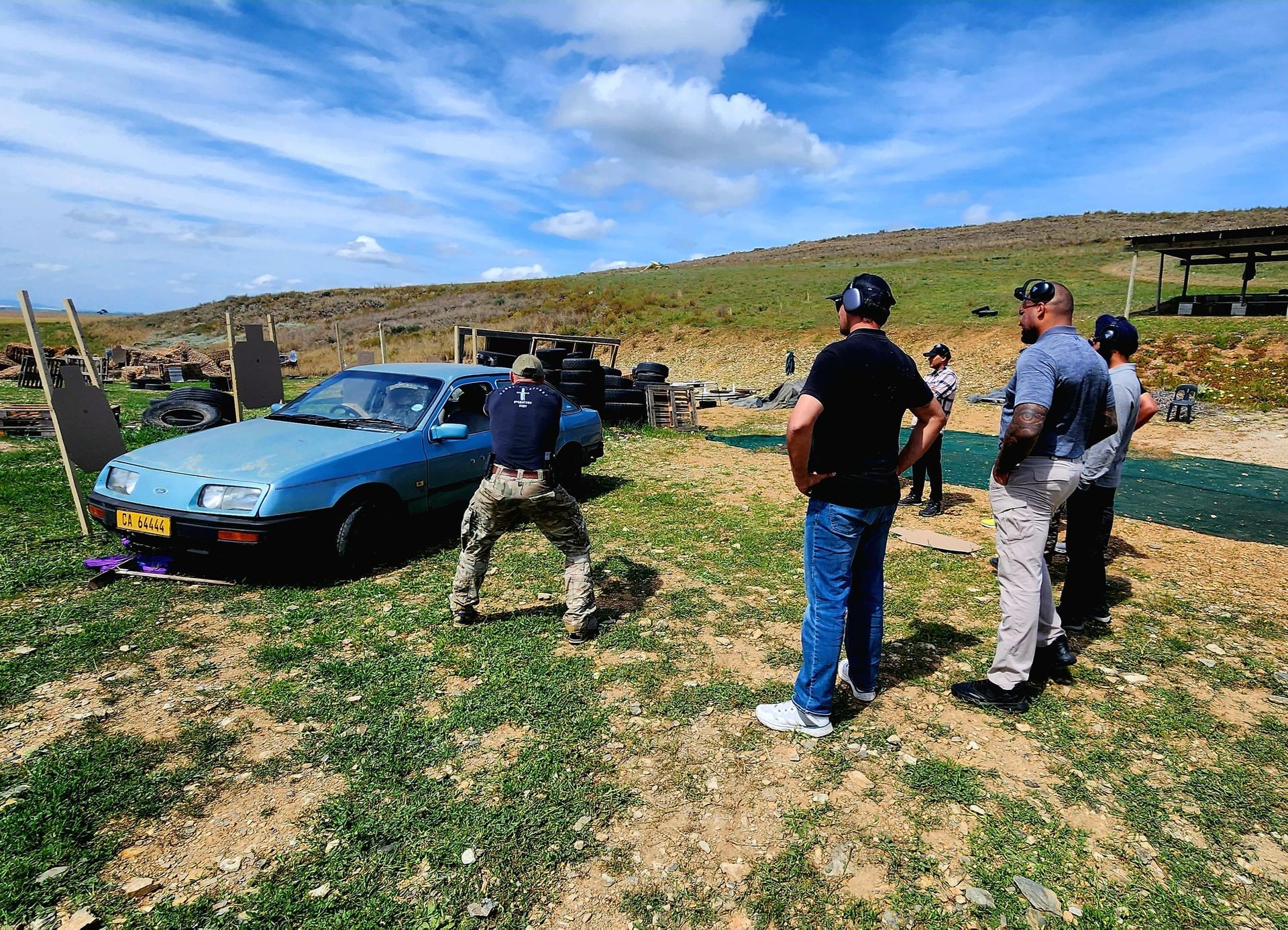 Group of people observing a man practicing shooting near an old blue car in an outdoor range.